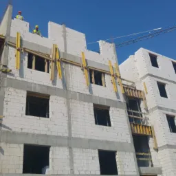 Wznoszenie wielopiętrowego budynku z białych bloczków, widoczne rusztowania i pracownicy w kaskach na górze, w tle błękitne niebo i żuraw budowlany.