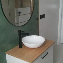 Biała umywalka nablatowa z czarną armaturą na szafce z bambusowym blatem, okrągłe lustro w czarnej ramie odbija kabinę prysznicową, ściany wyłożone płytkami w odcieniach zieleni i szarości.