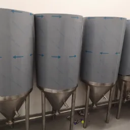 Cztery stalowe zbiorniki fermentacyjne na trzech nogach, z niebieską folią ochronną i strzałkami, stojące w pomieszczeniu z białymi ścianami i szarą podłogą.