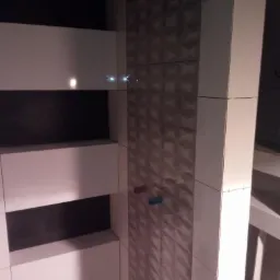 Ściana łazienki wyłożona białymi płytkami z poziomymi czarnymi pasami i kolumna z trójwymiarowymi płytkami w odcieniach szarości, widoczne przyłącza wodne i poziomica laserowa.