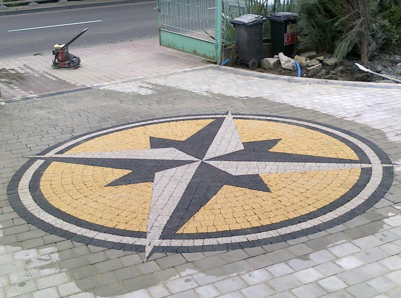 Ozdobny bruk z motywem rozety kompasowej, wykonany z kostki w odcieniach szarości, bieli i żółci. W tle widoczna zagęszczarka wibracyjna, brama i śmietniki.