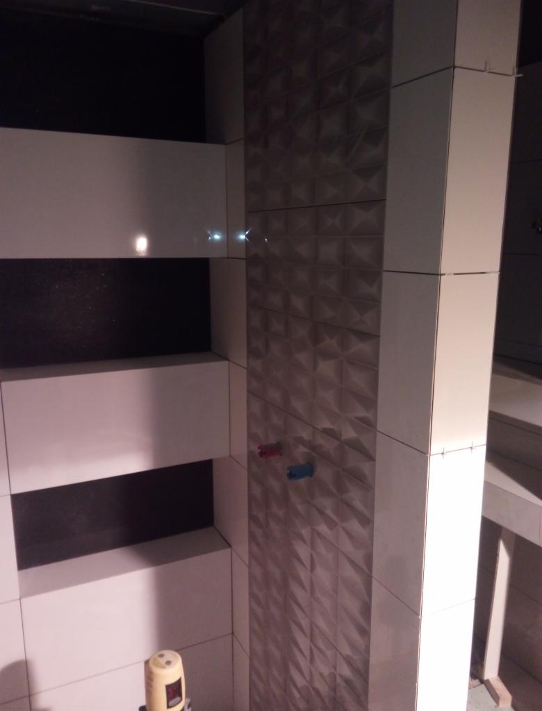 Ściana łazienki wyłożona białymi płytkami z poziomymi czarnymi pasami i kolumna z trójwymiarowymi płytkami w odcieniach szarości, widoczne przyłącza wodne i poziomica laserowa.