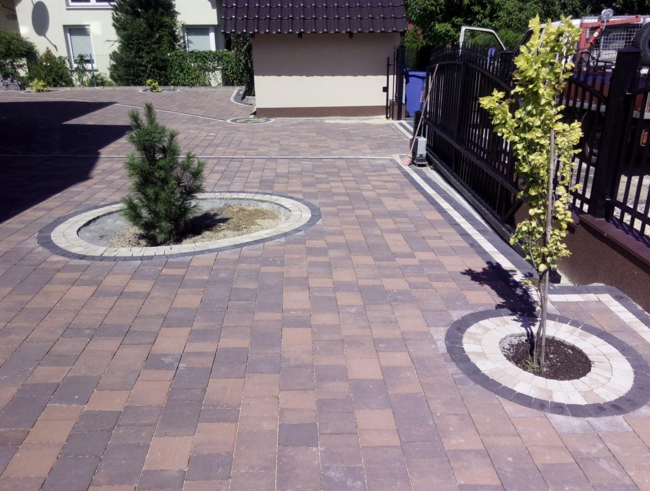 Ułożona kostka brukowa w dwóch odcieniach brązu, z okrągłymi obrzeżami wokół młodych drzewek i krzewów, prowadząca do bramy wjazdowej.
