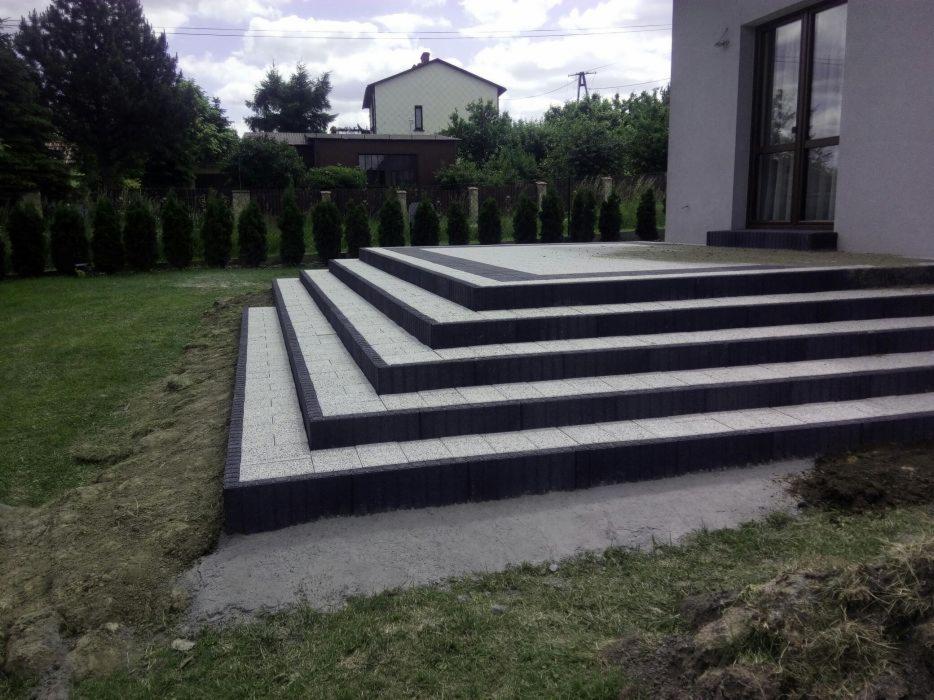 Schody tarasowe z jasnoszarej kostki brukowej z ciemnoszarą obwódką, prowadzące do domu, widoczne niedokończone prace ziemne.