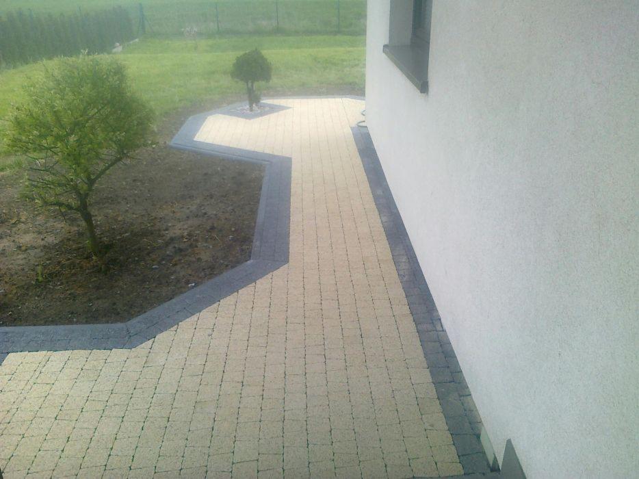 Ułożona ścieżka z jasnej kostki brukowej z ciemnoszarą obwódką prowadząca wzdłuż ściany domu, obok rabata z młodym drzewkiem.