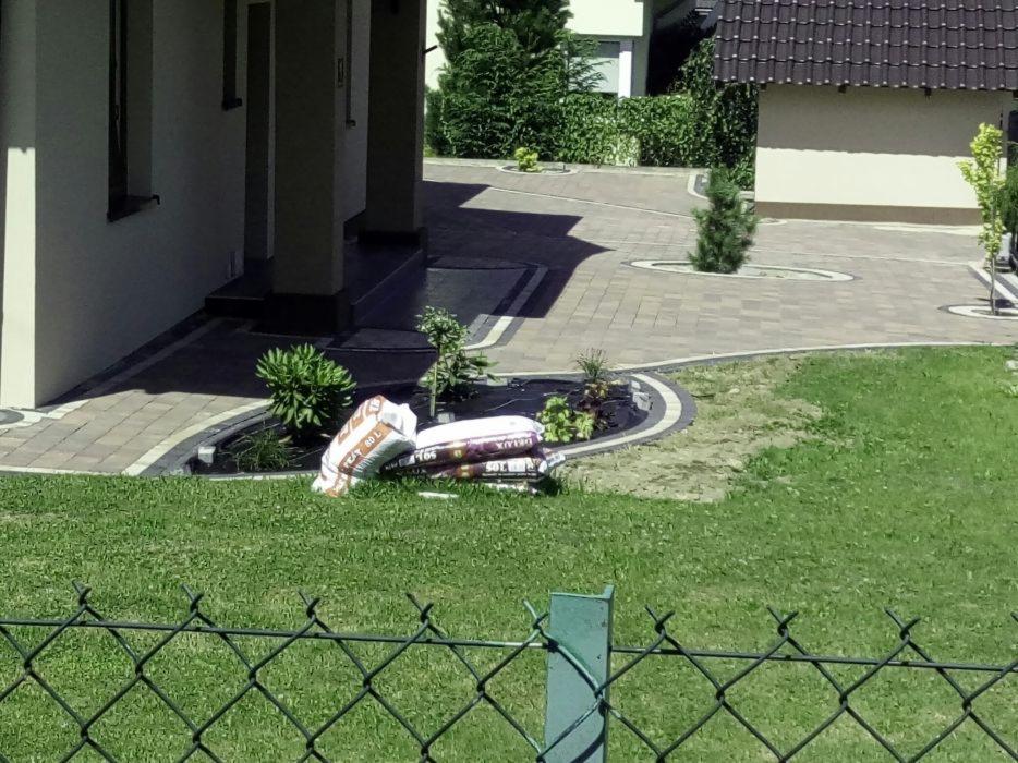 Ułożona kostka brukowa na podjeździe i ścieżce wokół domu, widoczne worki z ziemią ogrodową na trawniku, ogrodzenie z siatki w pierwszym planie.