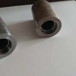 Dwa metalowe, cylindryczne elementy z gwintowanym otworem wewnętrznym, leżące na białym tle. Jeden element ma ślady obr&oacute;bki cieplnej lub korozji.