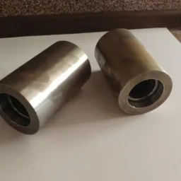 Dwa metalowe, cylindryczne elementy z otworem, leżące na białej powierzchni. Widoczne ślady obróbki i delikatne refleksy świetlne.