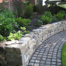 Ogród z kamienną rabatą i ścieżką z kostki granitowej, widok na zadaszone miejsce parkingowe w tle.