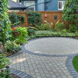 Okrągły taras z jasnoszarej kostki, obrzeże z ciemnoszarej kostki, otoczony bujną zielenią, drewniany płot w tle z dekoracją w kształcie słońca.