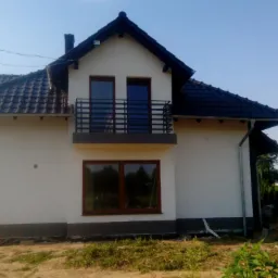 Dwupiętrowy dom z ciemną dachówką i balkonem, świeżo po malowaniu elewacji na biało, z widocznymi śladami po pracach wykończeniowych na trawniku.