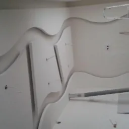 Wnętrze z dekoracyjnym sufitem podwieszanym z płyt kartonowo-gipsowych, o falistym kształcie i elementami pionowymi, w trakcie montażu, widoczne przewody i drabina w tle.