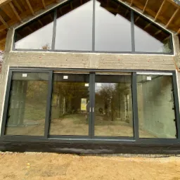 Nowoczesna fasada domu w stanie surowym z zamontowanymi dużymi oknami aluminiowymi w kolorze antracytowym, w tym przesuwne okno tarasowe i okno trójkątne pod dachem.