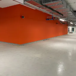 Pomarańczowa ściana w nowym biurze lub hali, szare wykończenie przy podłodze, widoczne instalacje pod sufitem, surowa podłoga z efektem połysku.