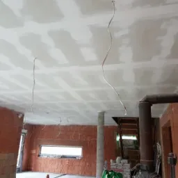 Wnętrze w trakcie prac wykończeniowych: sufit pokryty płytami gipsowo-kartonowymi, widoczne przewody elektryczne zwisające z sufitu, ściany z cegły, w tle piec kominkowy z rurą.