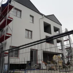 Nowo otynkowana elewacja budynku w trakcie wykańczania, widoczne rusztowania, siatka zabezpieczająca oraz maszyna do mieszania zaprawy, całość ujęta przez metalowe ogrodzenie.