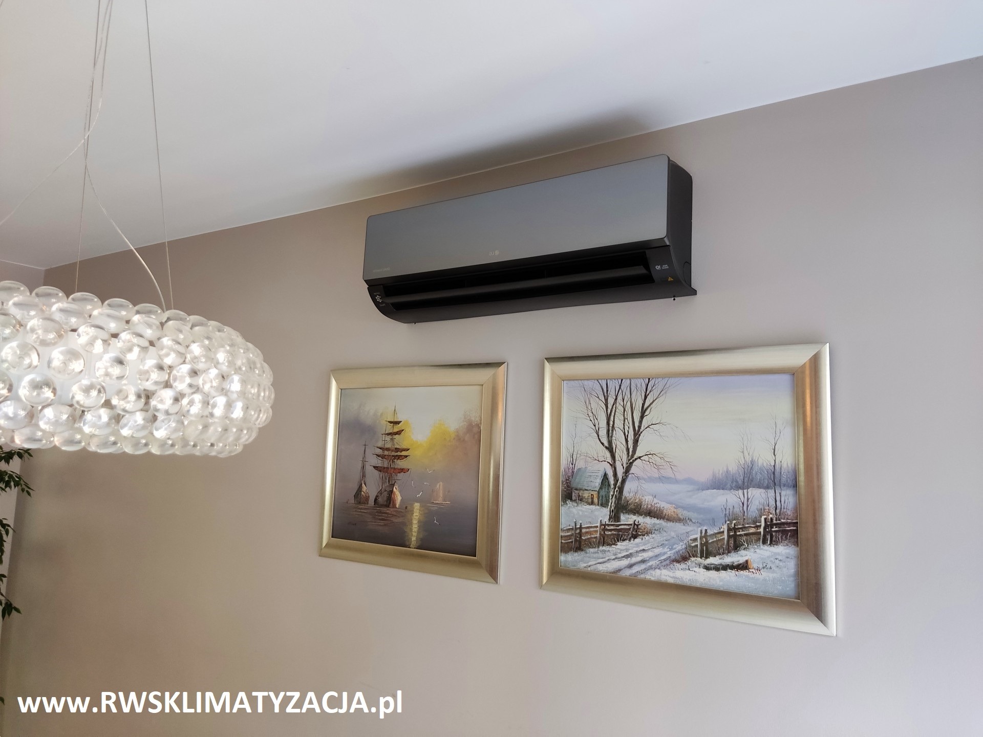 Czarny klimatyzator ścienny LG w nowoczesnym salonie z obrazami w srebrnych ramach i ozdobnym żyrandolem.