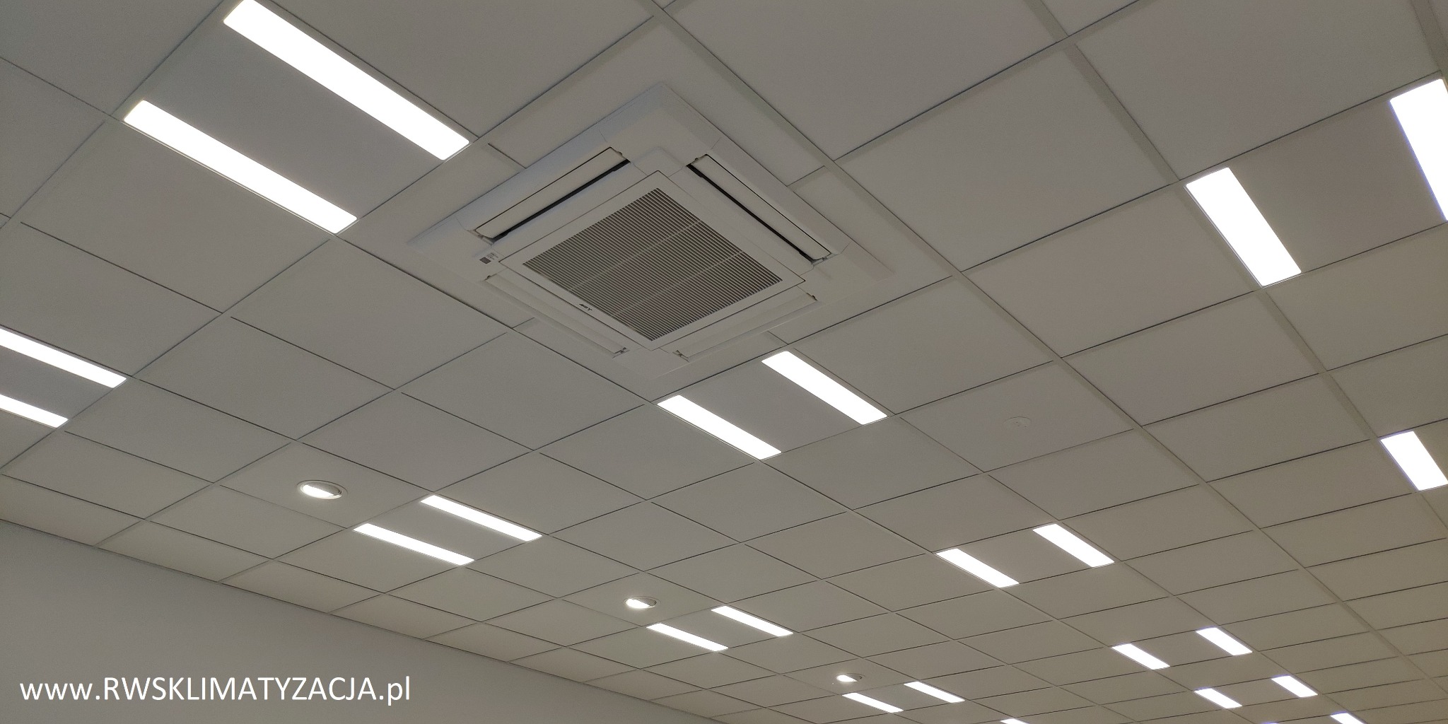 Kasetonowa sufitowa jednostka klimatyzacyjna w biurze z oświetleniem rastrowym, widok z dołu pod kątem, logo firmy w lewym dolnym rogu.