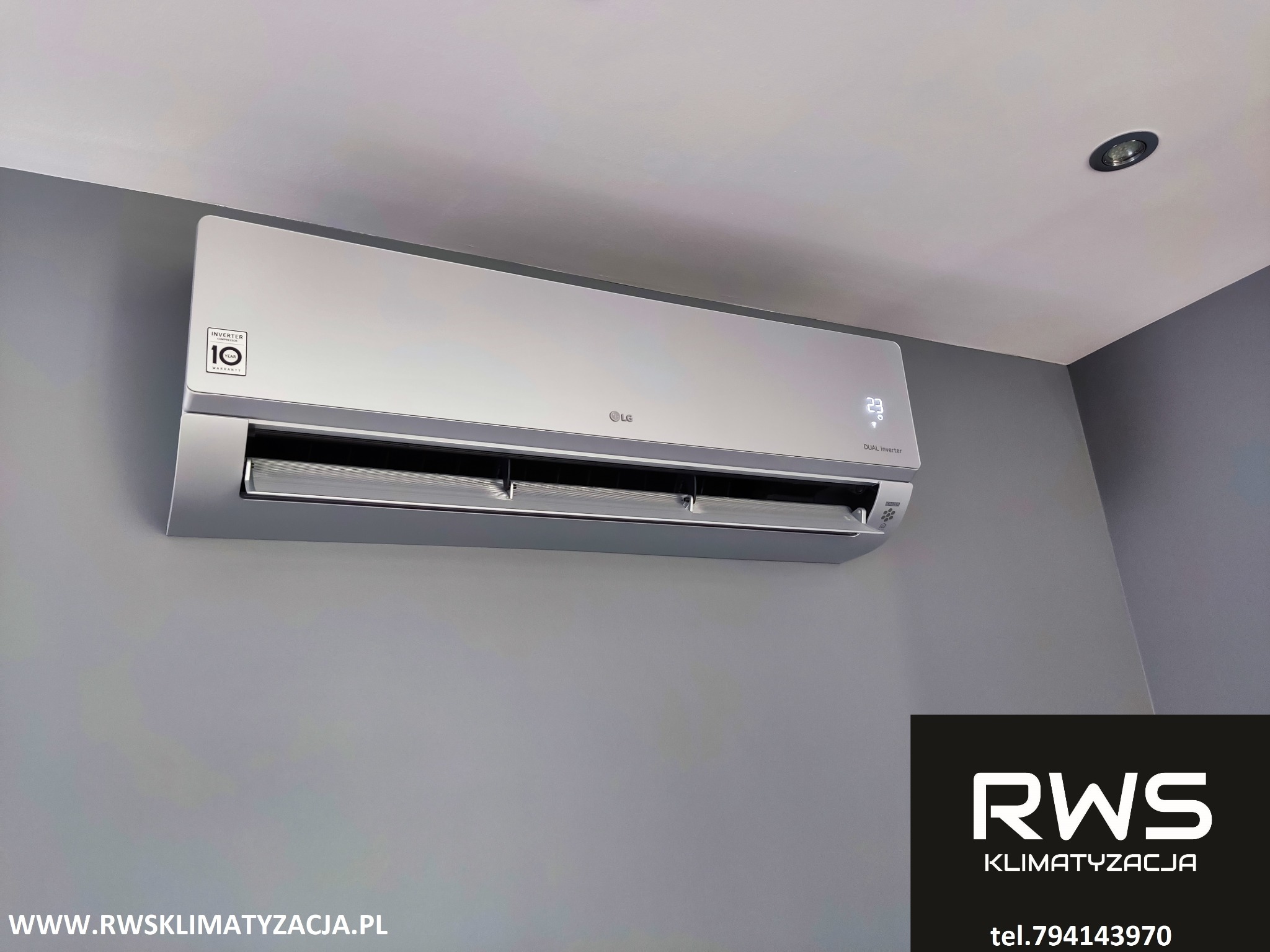 Zamontowany srebrny klimatyzator LG z wyświetlaczem temperatury 23 stopnie Celsjusza na szarej ścianie, widoczny fragment sufitu i logo RWS Klimatyzacja z adresem strony i numerem telefonu w prawym...