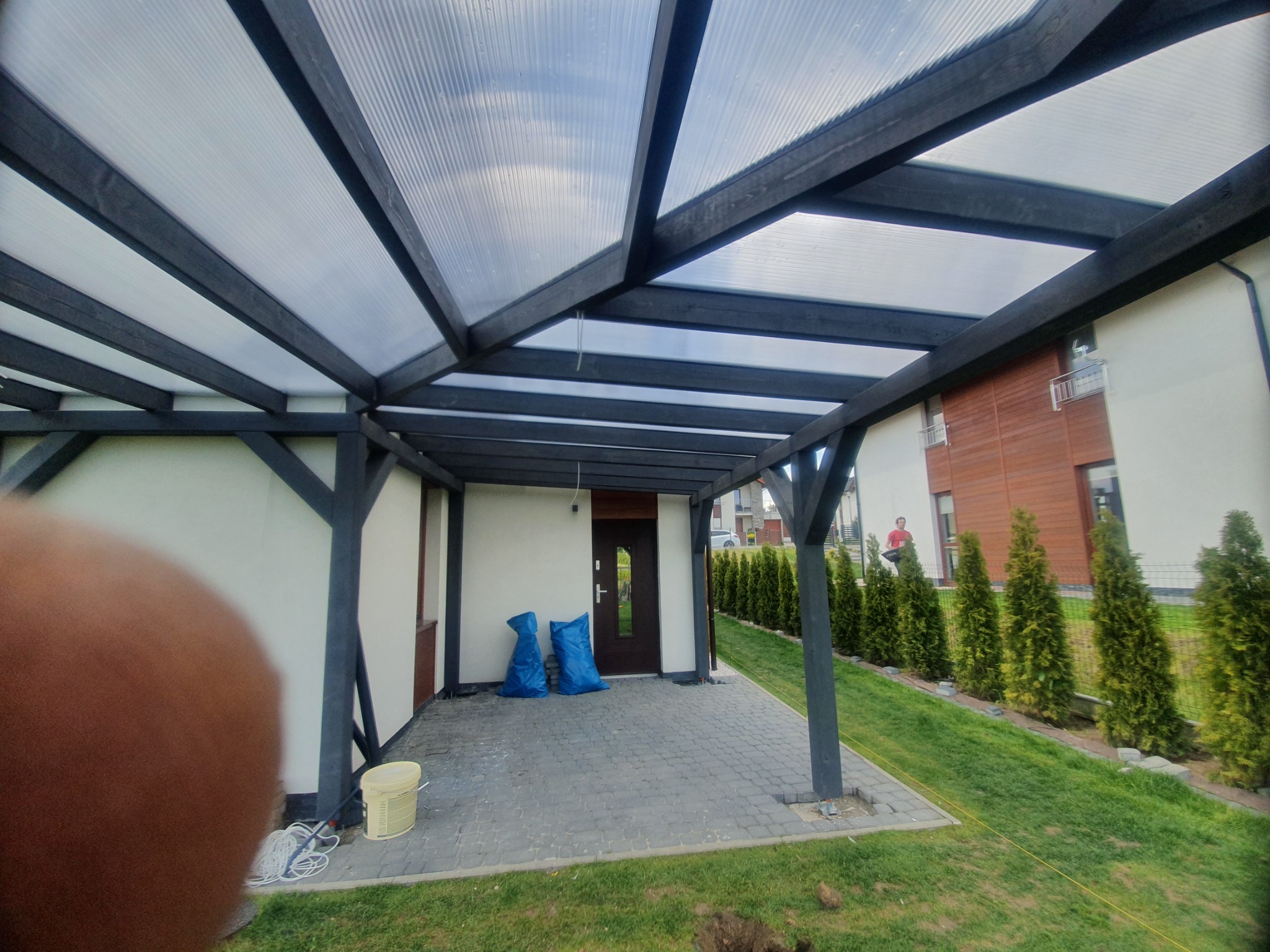 Zadaszenie tarasu z poliwęglanu i ciemnego drewna, widoczne dwa worki budowlane przy drzwiach wejściowych i częściowo przesłonięte ujęcie