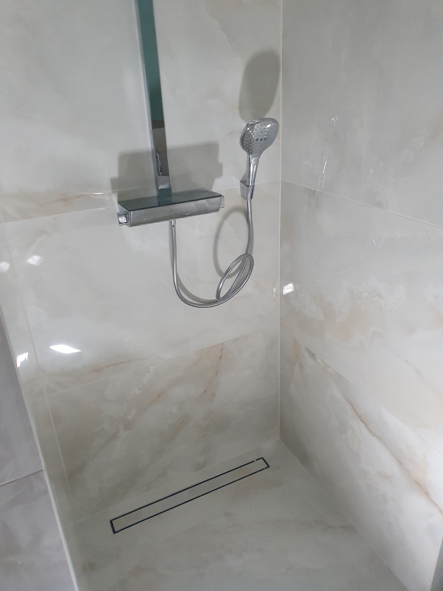 Nowoczesny prysznic z chromowaną armaturą i odpływem liniowym, wykończony płytkami imitującymi marmur.