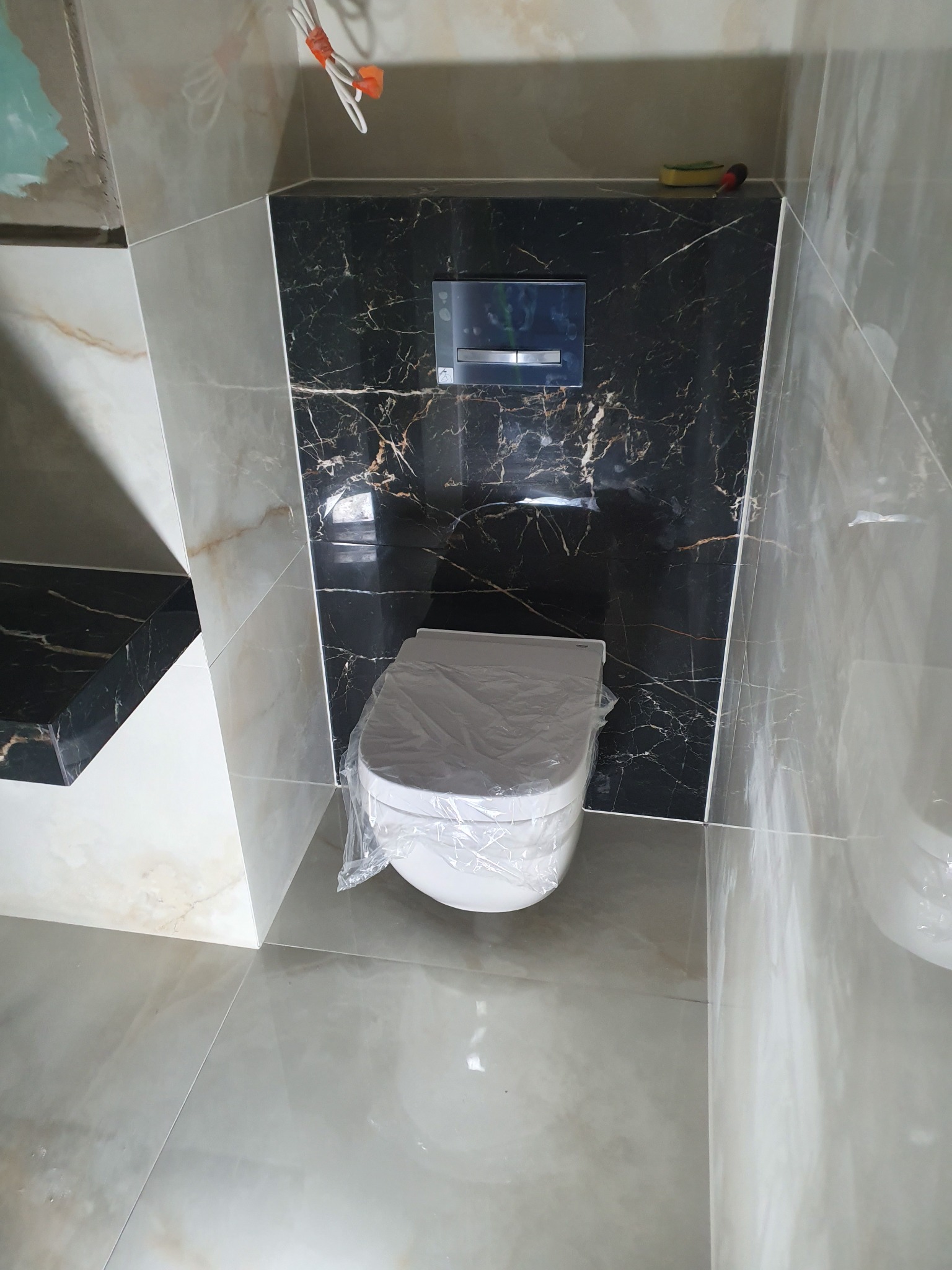 Nowa toaleta wisząca, jeszcze w folii ochronnej, zamontowana w wykończonej łazience z płytkami imitującymi marmur w odcieniach bieli, szarości i czerni z wyraźnym użyleniem.