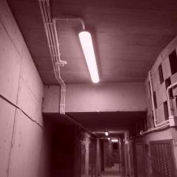 GLOBAL Tomasz Zatyka - Korytarz piwniczny z widocznymi rurami instalacji elektrycznej pod sufitem i lampą jarzeniową, betonowe ściany z otworami wentylacyjnymi.