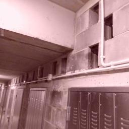 GLOBAL Tomasz Zatyka - Korytarz piwniczny z rzędami metalowych szafek, widoczne instalacje rurowe pod sufitem i ściany z betonowych pustaków.