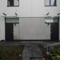 GLOBAL Tomasz Zatyka - Dwa wejścia do budynku z nowoczesnymi szklanymi zadaszeniami wspartymi na metalowych wspornikach, widoczne czarne drzwi i jasna elewacja z płyt, brukowana ścieżka prowadząca do wejść, teren wokół...