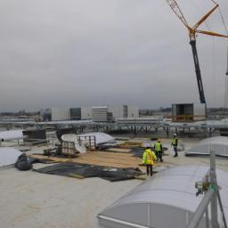 GLOBAL Tomasz Zatyka - Ekipa budowlana w kamizelkach odblaskowych na płaskim dachu, z dźwigiem w tle, montuje elementy konstrukcyjne; widoczne świetliki dachowe i elementy wentylacyjne.