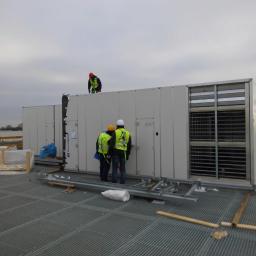 GLOBAL Tomasz Zatyka - Trzech pracowników w kamizelkach odblaskowych instaluje dużą centralę wentylacyjną na dachu budynku przemysłowego, widoczny metalowy stelaż i kratownica podłoża.