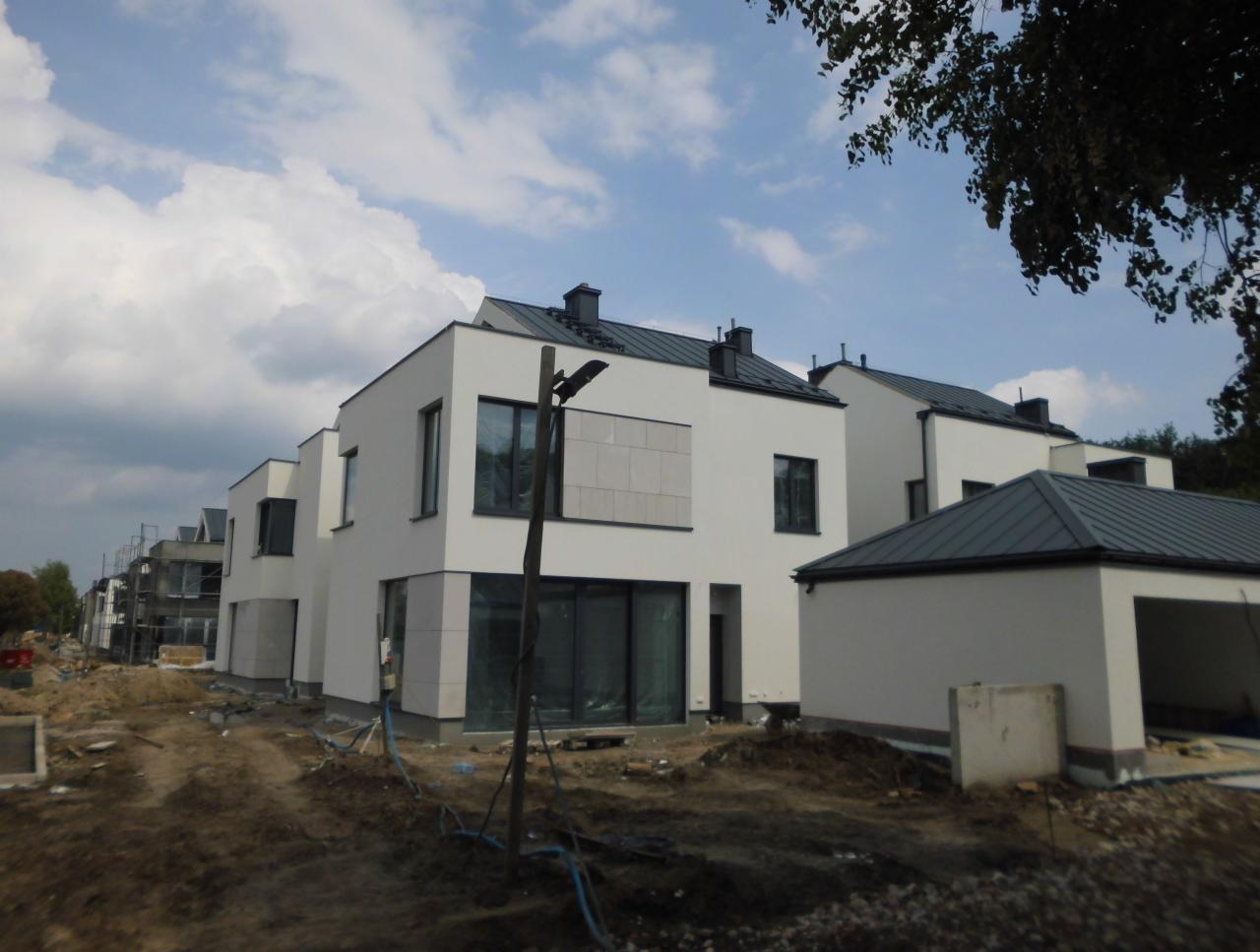 Nowoczesne, białe domy szeregowe z ciemnymi dachami w trakcie budowy, widoczne rusztowania i teren budowy z ziemią.