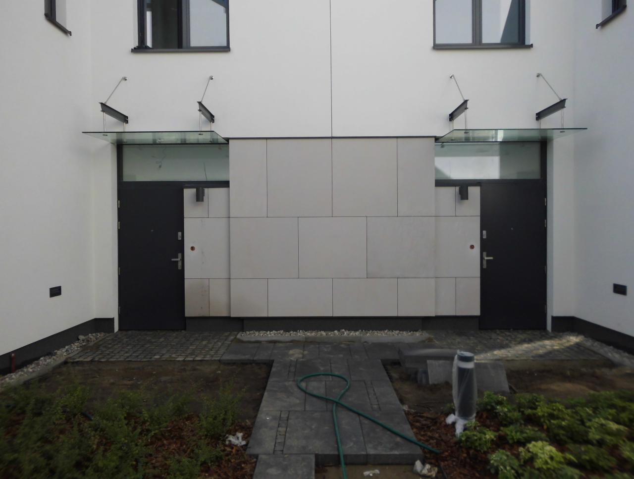 Dwa wejścia do budynku z nowoczesnymi szklanymi zadaszeniami wspartymi na metalowych wspornikach, widoczne czarne drzwi i jasna elewacja z płyt, brukowana ścieżka prowadząca do wejść, teren wokół...