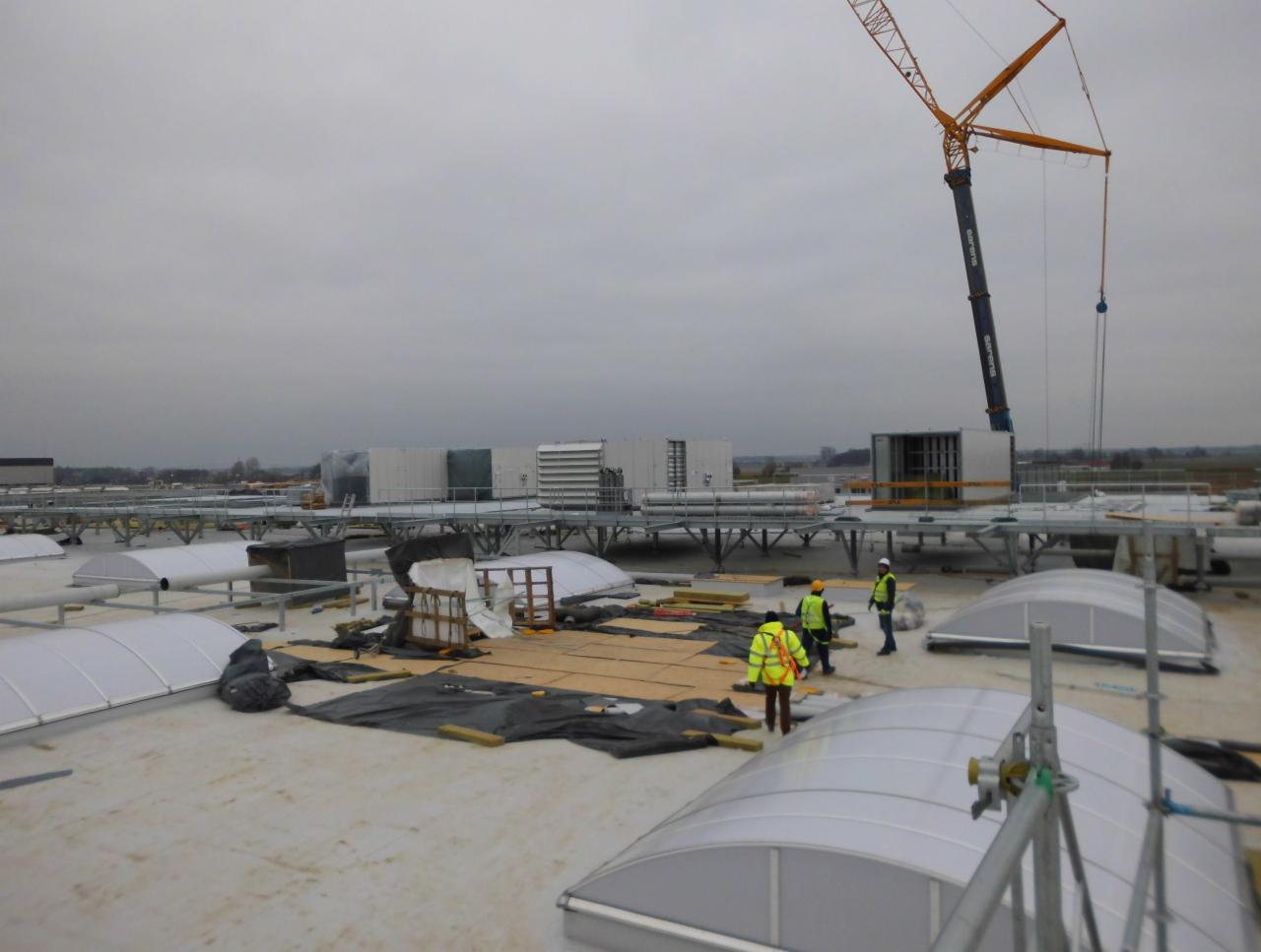 Ekipa budowlana w kamizelkach odblaskowych na płaskim dachu, z dźwigiem w tle, montuje elementy konstrukcyjne; widoczne świetliki dachowe i elementy wentylacyjne.