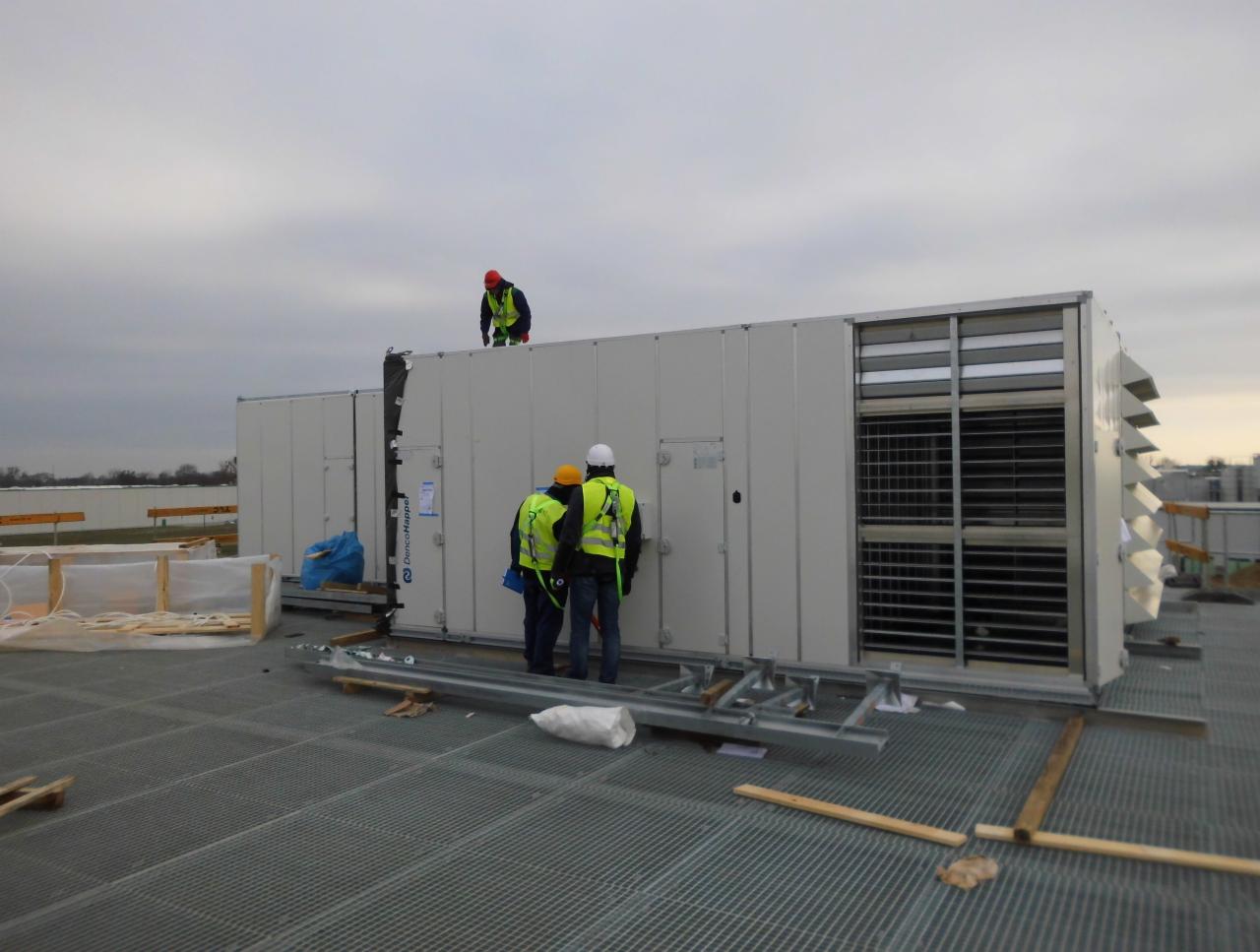 Trzech pracowników w kamizelkach odblaskowych instaluje dużą centralę wentylacyjną na dachu budynku przemysłowego, widoczny metalowy stelaż i kratownica podłoża.