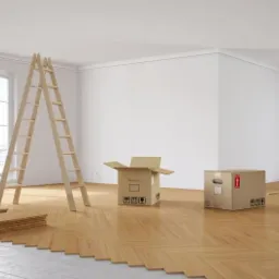 Parkiet ułożony w jodełkę w jasnym pokoju, częściowo przykryty płytami podkładowymi, drabina oparta o ścianę, kartony, puszka z czerwoną farbą i wałek malarski w tle, widok na miasto za oknem.