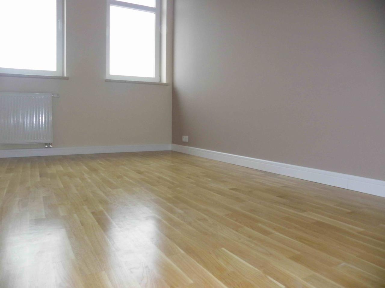 Pomalowane na beżowo wnętrze pokoju z drewnianą podłogą, białymi listwami przypodłogowymi, grzejnikiem pod oknem i widocznym gniazdkiem elektrycznym.