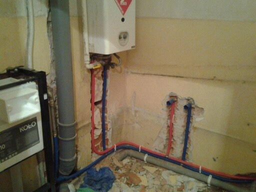 Wewnętrzne rury wodne (czerwona i niebieska) wystające ze ściany obok szarej rury kanalizacyjnej i podgrzewacza wody, fragmenty gruzu na podłodze.