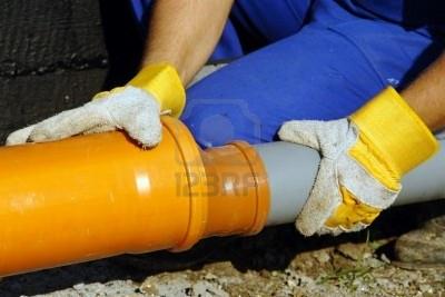 Instalator łączy pomarańczową rurę kanalizacyjną z szarą rurą, używając złączki; widoczne rękawice robocze z żółtymi mankietami i niebieski kombinezon.