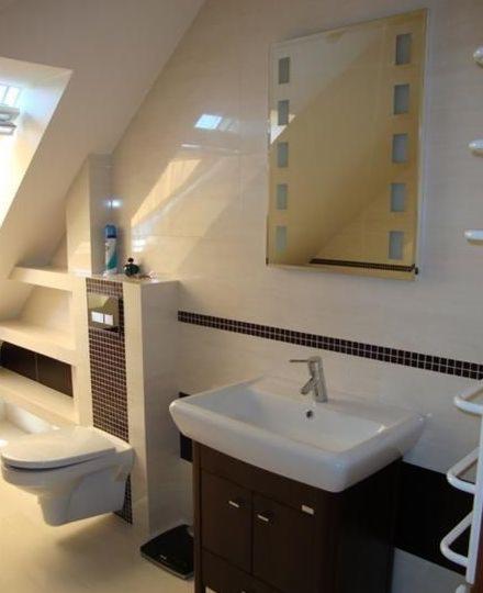 Biała łazienka na poddaszu z toaletą, umywalką na szafce w kolorze ciemnego drewna, lustrem z dekoracyjnymi wstawkami i czarnym paskiem mozaiki na ścianie.