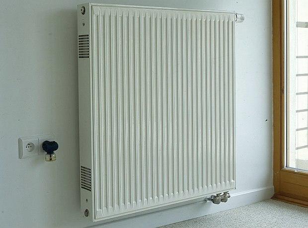 Biały grzejnik panelowy zamontowany na białej ścianie, widoczne przyłącza wodne i gniazdko elektryczne z termostatem.