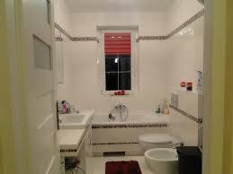 Jasna łazienka z białą armaturą, wanną obudowaną płytkami z dekoracyjnym pasem, umywalką z blatem i oknem z czerwoną roletą.