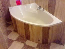 Narożna wanna z obudową z jasnego drewna o pionowym układzie desek, chromowana bateria, płytki podłogowe w szachownicę, fragment ściany wyłożonej płytkami w odcieniach beżu.