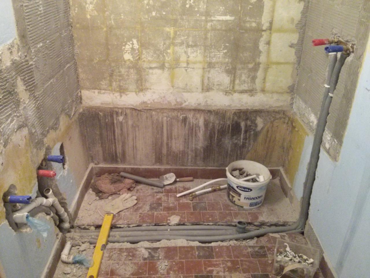 Remont łazienki w trakcie: przygotowane ściany do położenia płytek, rury instalacji wodnej z czerwonymi i niebieskimi zaślepkami, poziomica, wiadro z resztkami zaprawy i narzędzia na podłodze...