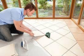 Mężczyzna w trakcie układania jasnych płytek podłogowych w oranżerii, używając poziomicy krzyżowej dla precyzyjnego ułożenia.