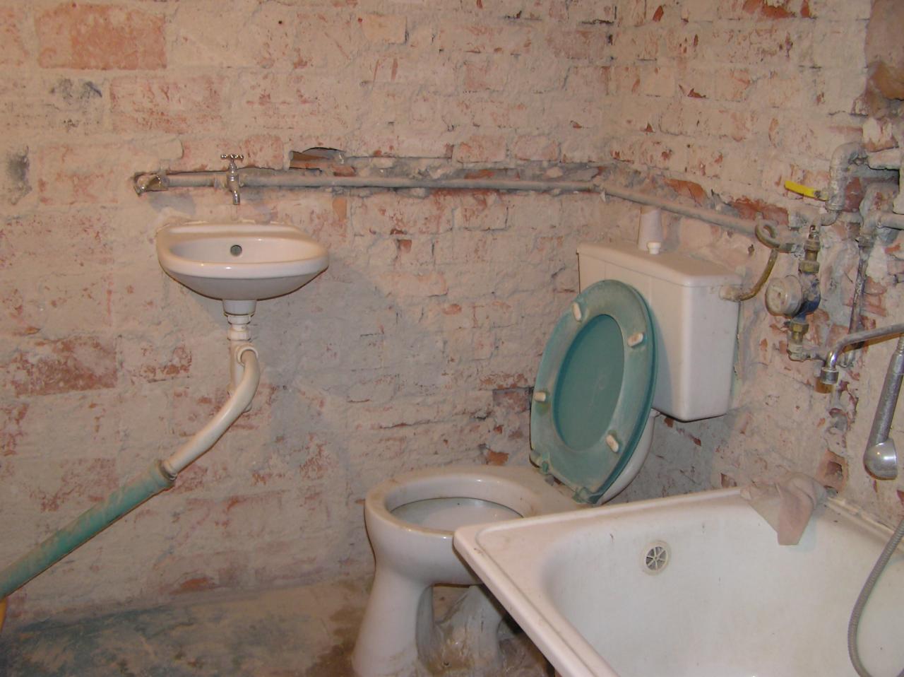 Łazienka w trakcie remontu z odsłoniętą cegłą, widoczna umywalka, toaleta z podniesioną deską w kolorze morskim i fragment wanny.