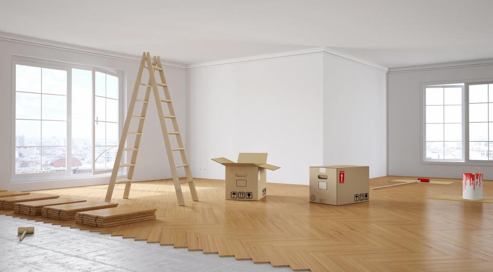 Parkiet ułożony w jodełkę w jasnym pokoju, częściowo przykryty płytami podkładowymi, drabina oparta o ścianę, kartony, puszka z czerwoną farbą i wałek malarski w tle, widok na miasto za oknem.