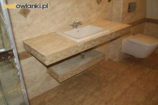 Wykończona łazienka z beżowymi płytkami, umywalką nablatową na blacie z dekoracyjnym obramowaniem i podwieszaną toaletą.