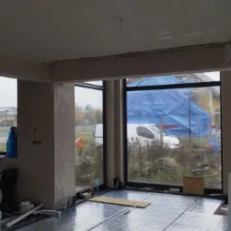 Wnętrze w trakcie prac wykończeniowych: ściany po nałożeniu tynku, instalacja ogrzewania podłogowego na folii izolacyjnej, widok na zewnątrz przez duże okna z budynkiem osłoniętym niebieską plandeką.