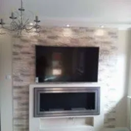 Ściana telewizyjna z dekoracyjną okładziną imitującą kamień, z wbudowanym biokominkiem w metalowej obudowie i minimalistycznym oświetleniem punktowym, zwieńczona nowoczesnym telewizorem LCD.